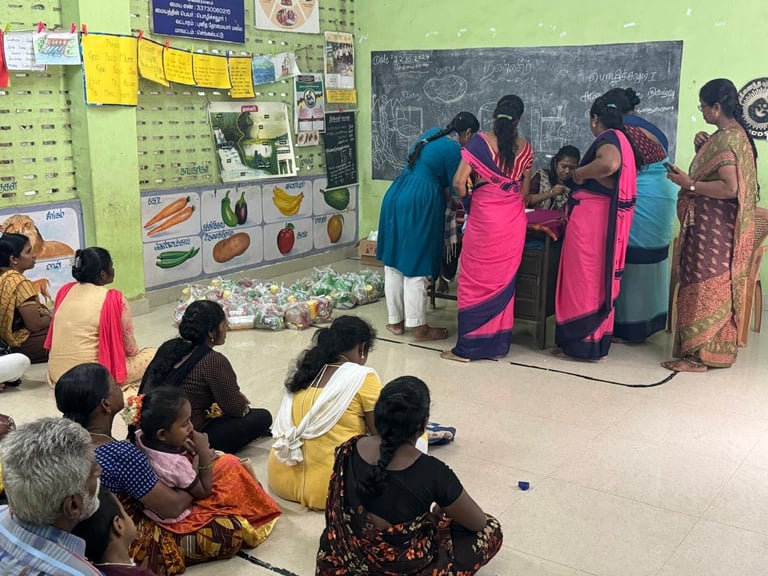 Women and children in colorful clothing gathered in a classroom with a green wall, some seated on the floor and others standing near a chalkboard during a community education session