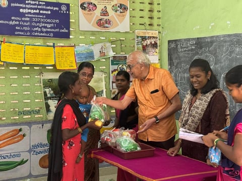 An elderly man in an orange shirt distributes fresh vegetables to women in a classroom setting with educational posters on the walls