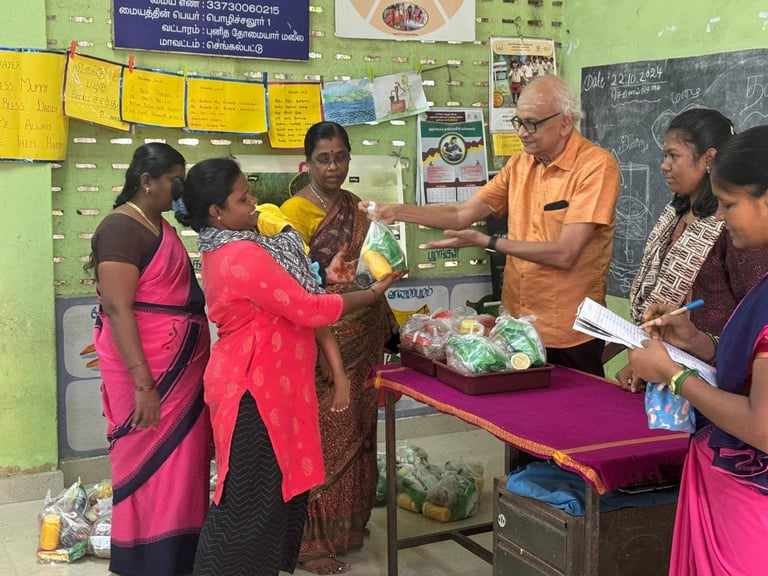 Teacher in orange shirt demonstrating produce to female students wearing traditional Indian attire in a classroom with green walls