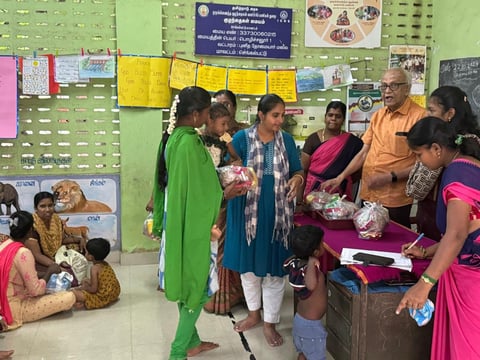 Group of people gathered in a community center or classroom with green walls, receiving or distributing food items from a table