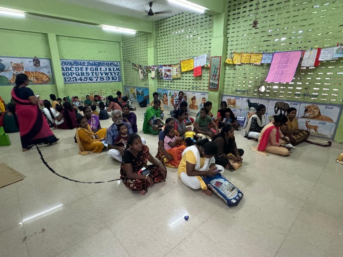 Group of children sitting on floor in classroom during learning activity with colorful posters on green walls